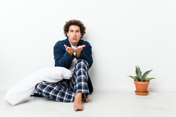 Mature man wearing pajama sitting on house floor folding lips and holding palms to send air kiss.