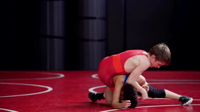 Youth wrestlers in red and blue singlets live wrestling from the top and bottom position. Red throws his legs in on the blue wrestler.