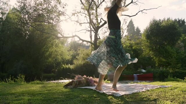 Young woman swirling around in park during picnic with sun shining through trees, dress is swaying around in air, hipster girl with straw hat, slow motion