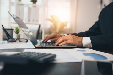 Close up hand of business man typing and working on laptop with  finance graph paper on table,...