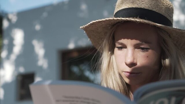Close up of a young woman with a straw hat on reading a book at sunset on a rural farm in Ibiza, Spain. Circling shot in slow motion.