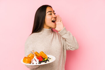 Young asian woman holding a waffle isolated shouting and holding palm near opened mouth.