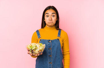 Young asian woman holding a salad isolated shrugs shoulders and open eyes confused.