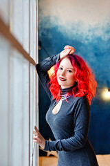 A young beautiful woman with bright red hair at home is smiling standing at the window, looking at the camera. Positive people.