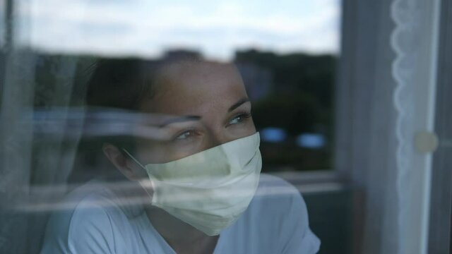 Young woman wears a mask to protect herself from covid-19 while looking out the window. slow motion