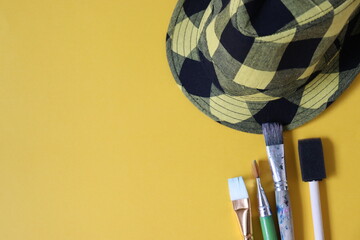 Yellow background with paint brush and plaid hat.
