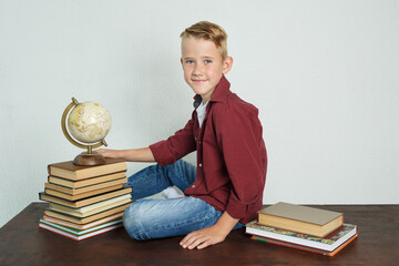 A schoolboy sits at a desk near books and holds a globe with his hand.