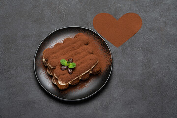 portion of Classic tiramisu dessert on ceramic plate and heart shaped cocoa powder on concrete...