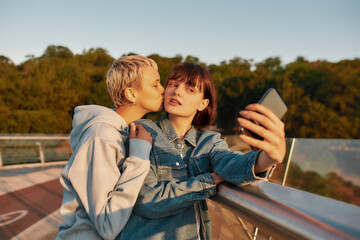 Lesbian couple standing on the bridge and posing while taking a selfie picture together