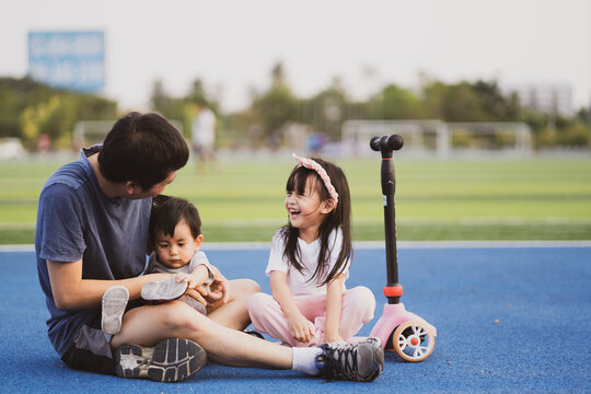 Asian Father And Child Sitting In Stadium After Exercise Time, They Relaxing Talk And Laughing Together With Happiness Moment, Concept Of Sport And Outdoor Activity, Quality Time Spending In Family.