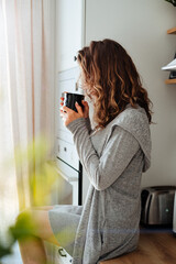 Young pretty woman drinks coffee sitting at her kitchen at home and looking out the window enjoys of morning.