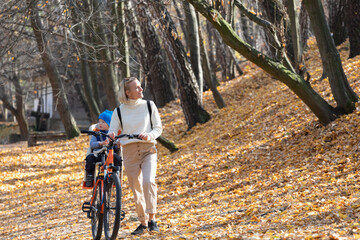 Happy mother leads a bicycle with a child strapped in the back in the autumn park.