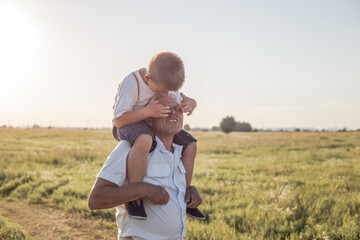 Fototapeta premium Portrait of cute little boy and her handsome grandpa looking at camera and smiling on meadow. kid is sitting pickaback