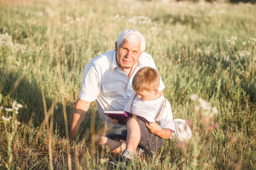 Naklejka na ściany i meble Mid shot of grandfather and his grandson while reading a book together in meadow Small boy making first steps in read.