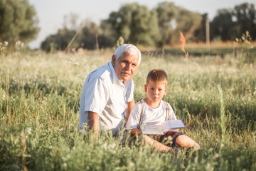 Naklejka na ściany i meble Mid shot of grandfather and his grandson while reading a book together in meadow Small boy making first steps in read.