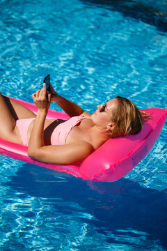 Sexy woman with a phone in a swimsuit lies on a pink inflatable mattress in the pool. Relax by the pool on a hot summer sunny day. Vacation concept