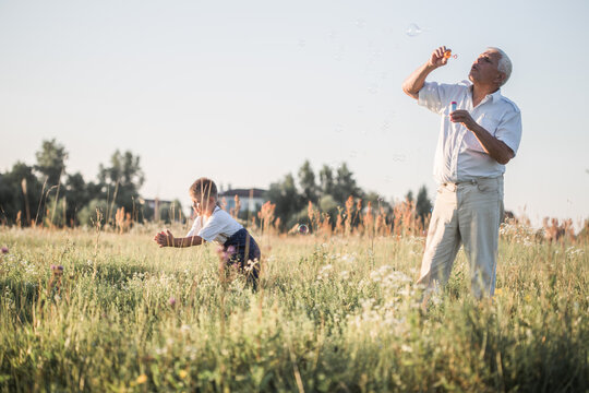 Happy senior man Grandfather with cute little boy grandson playing in field.  Male blowing soap bubbles while an excited kid enjoys the bubbles. Happy teenage