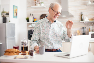 Cheerful senior man on online video call from kitchen while enjoying breakfast and a cup of coffee. Elderly person using internet online chat technology video webcam making a video call connection