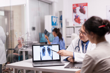 Mature doctor pointing at patient lungs ct scan on laptop during medical visit in hospital office....