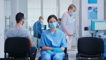 Obraz premium Worried nurse with face mask against coronavirus sitting on chairs in hospital waiting room. Disabled senior woman with walking frame in hospital hallway.