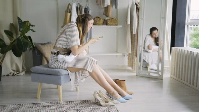 elegant long haired lady in striped dress holds little baby in arms and looks at photos of new shoes on smartphone sitting on designed chair at home