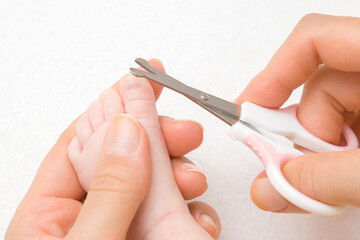 Young mother hand holding infant foot and cutting toenail with baby scissors on light gray background. Closeup. 