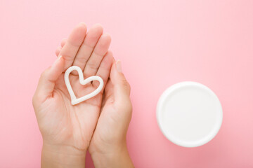 Heart shape on young woman palm. White cream jar on light pink table background. Pastel color. Care...