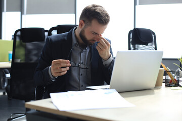 Fototapeta premium Handsome young business man with closed eyes touching face with hands while sitting on working place in office.