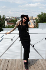 Happy smiling woman in black dress, hat and sunglasses standing on rooftop outdoors, copy space