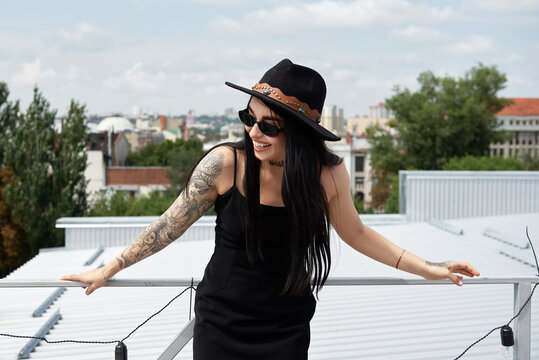 Happy smiling woman in black dress, hat and sunglasses standing on rooftop outdoors, copy space