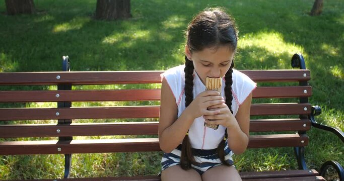 Pretty Caucasian  girl with long braids eating a french hot dog on a park bench. Portrait of a girl in a white T-shirt who eats fast food. 4k 50 fps slow motion
