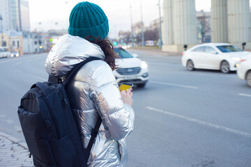 girl in a down jacket and hat stands at a public transport stop and uses a smartphone. Wait for the bus.