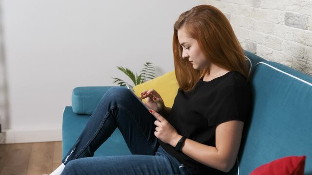 Young girl is sitting on a blue sofa and watching the news using a tablet. Serious and concentrated girl.