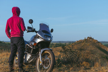 Obraz premium Back view, biker in a red sweatshirt with a hood. Motorcycle driver at the top of the hill overlooking the rocks and the river. Hobbies and leisure. Far from civilization.