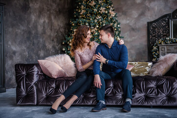 A fashion couple in love sits on a modern sofa in the middle of a dark loft. In the background there is a festive tree decorated with a garland of lights, golden balls, a vintage fireplace with gifts.