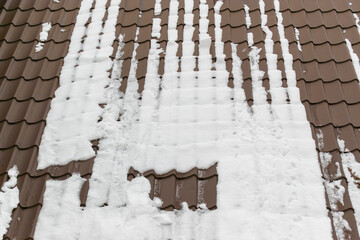 The roof is made of metal tiles, covered with white snow. Brown profiled metal sheet. Concept of the first snow in the winter season, the onset of cold weather.