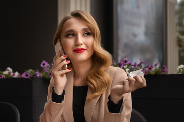 Charming young woman with long blonde hair is responding incoming phone call while sitting in a cafe, drinking coffee