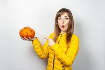 young girl with a surprised face holds a pumpkin and points a finger at it on a light background. Halloween concept, autumn, celebration