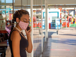 Mujer joven hablando por el teléfono móvil mientras viste mascarilla para cumplir las medidas de seguridad y distanciamiento de la pandemia del coronavirus en un centro comercial
