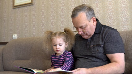 Granddaughter is reading a book with grandfather. The girl frowns at the book and listens carefully to Grandfather. Sitting on the couch together closeup