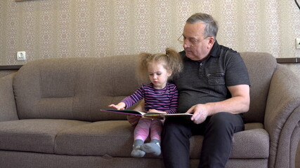 Granddaughter is reading a book with grandfather. The girl frowns at the book and listens carefully to Grandfather. Sitting on the couch together.