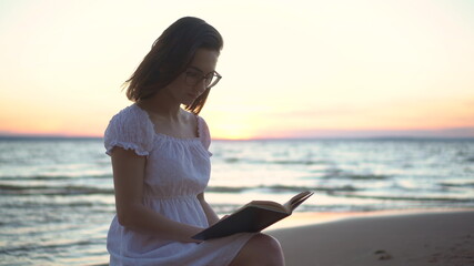 Fototapeta na wymiar A young woman sits on a stone on the beach by the sea with a book in her hands. A girl in a white dress at sunset reads a book closeup.