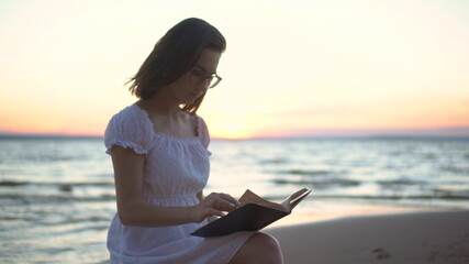 Fototapeta na wymiar A young woman sits on a stone on the beach by the sea with a book in her hands. A girl in a white dress at sunset reads a book closeup.