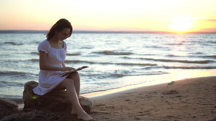 Fototapeta na wymiar A young woman sits on a stone on the beach by the sea with a book in her hands. A girl in a white dress at sunset reads a book.