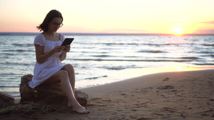 Fototapeta na wymiar A young woman sits on a stone on the beach by the sea with a tablet in her hands. A girl in a white dress at sunset is chatting on a tablet.