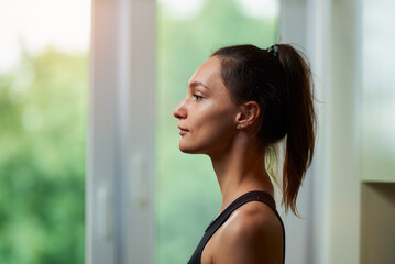 A side view photo of a sporty slim girl in a black workout tight suit is relaxing before training near the window at home.