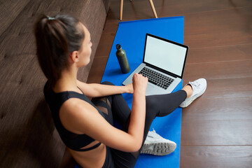 A sideways photo of a sporty girl in black tight pants and top is sitting on the blue yoga mat with a computer at home. A mockup of a laptop and fitness woman.