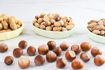 Hazelnuts in shells lie on a white table. Behind him are three bowls of shelled nuts (cashews, pistachios, almonds). Healthy food high in antioxidants, protein and vitamins. Close-up