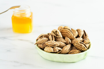 Salted almonds in shells in a light green bowl stands on a white background. In the background is a small glass jar filled with honey and a spoon. Nuts go well with honey. Close-up