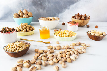 A lot of pistachio nuts are scattered on a white table. Wooden and ceramic bowls contain hazelnuts,...
